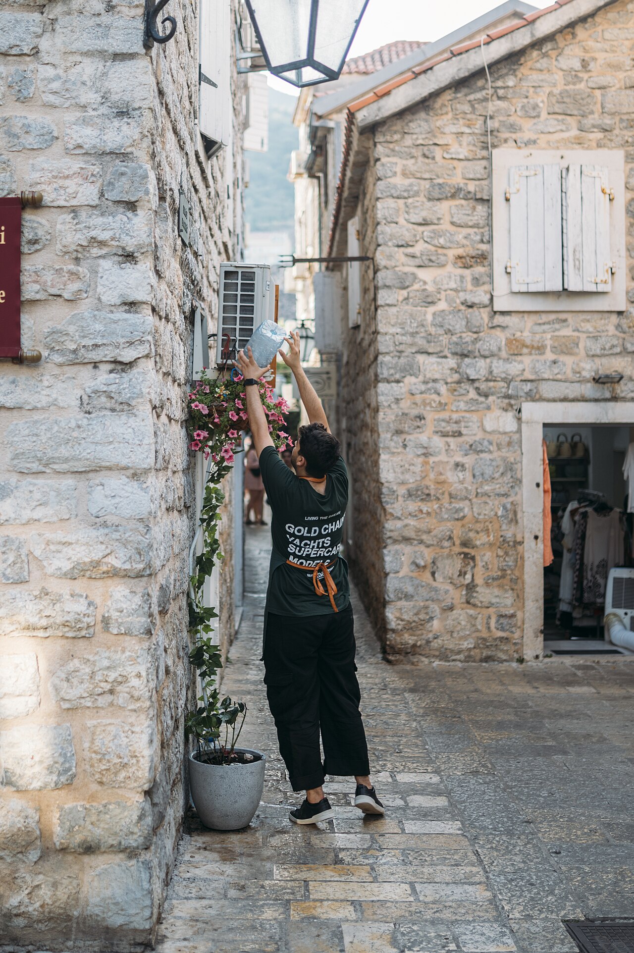 Person watering flowers in a narrow street lined with stone buildings in a Budva old town
